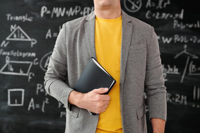 Close-up of a teacher holding a notebook in a classroom setting with equations on the blackboard.