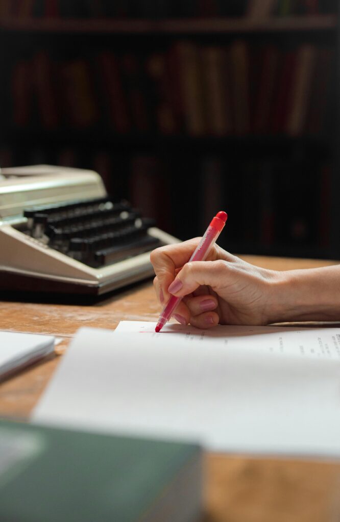 A close-up shot of a hand writing on papers with a red pen beside a vintage typewriter.